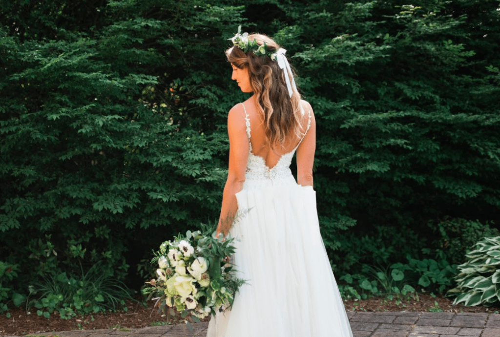 The back of a wedding dress with the bride wearing a flower crown and holding her bouquet down at her side.