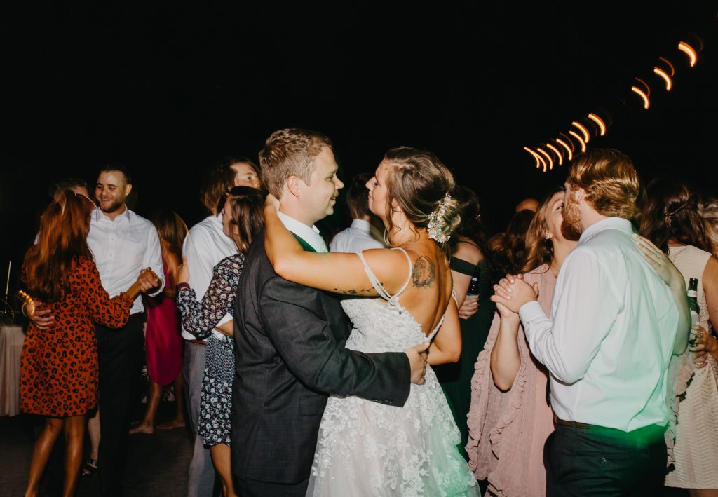 Bride and groom dancing together under bistro lights with their guests.