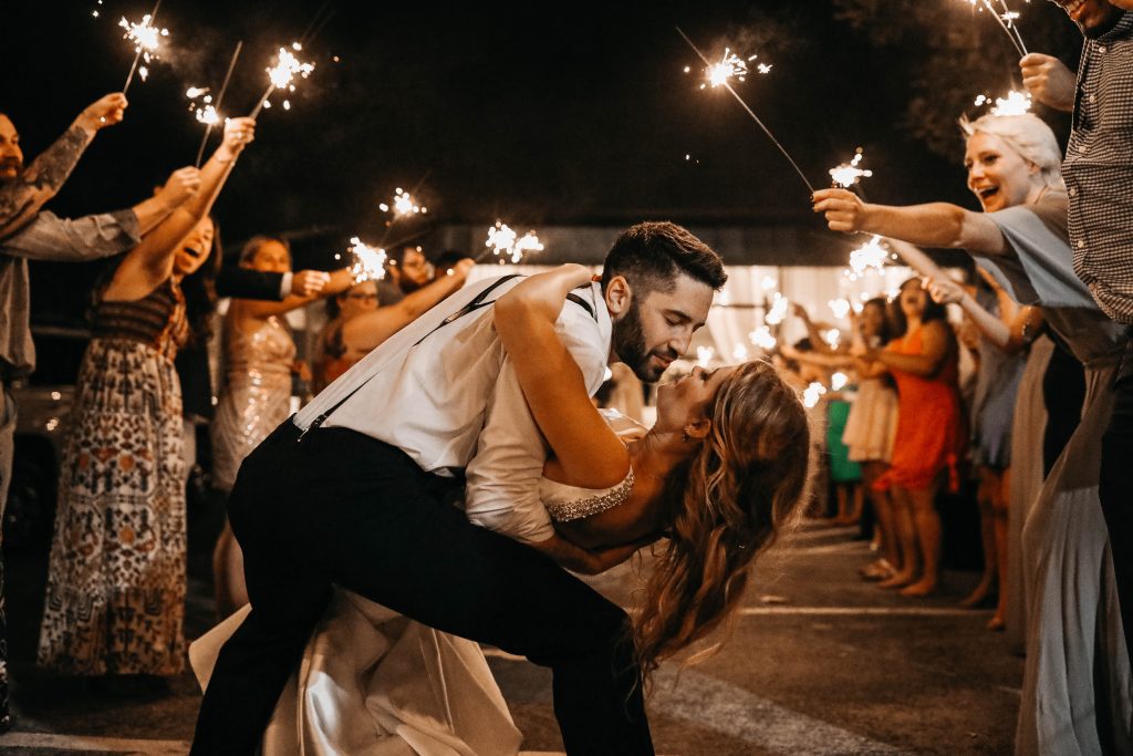 The groom dips his bride for a kiss with sparklers glowing in the background.