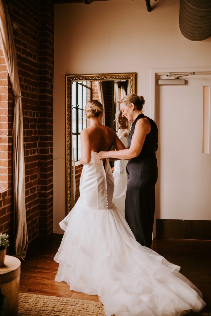 Mom helps her daughter get her wedding dress on in the bridal suite at the industrial chic venue.