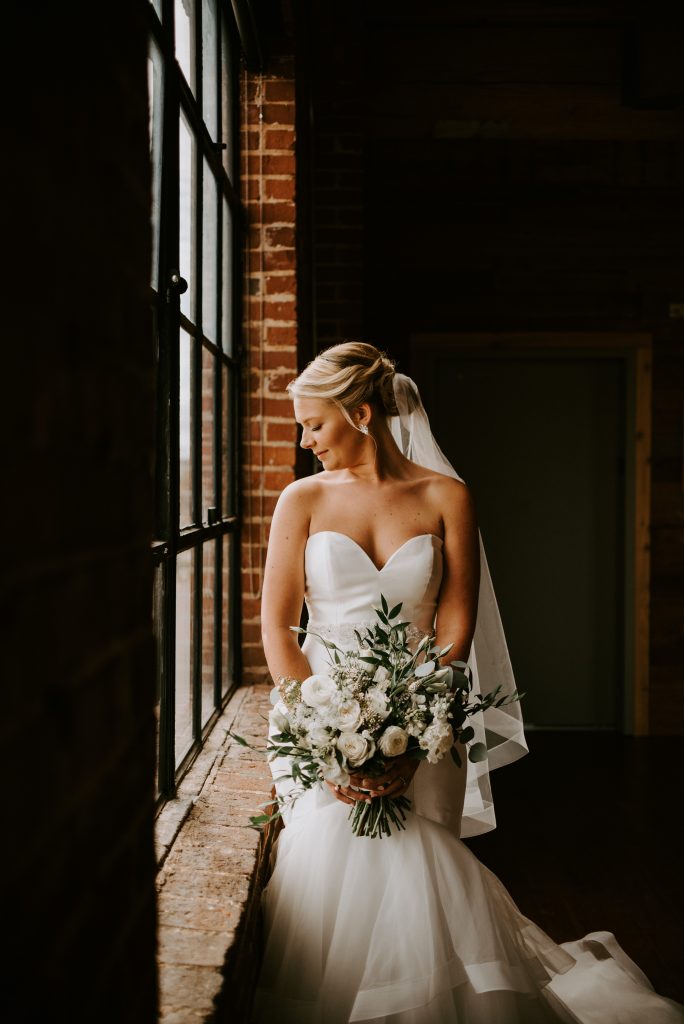 A stunning bridal portrait with the chic bride in front of industrial windows holding her bouquet and looking down. 